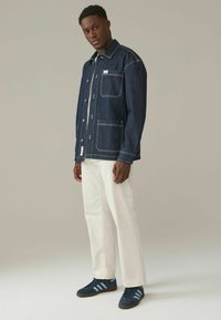 Jeansjakke med kontrasterende hvide syninger, to forlommer og et logo-patch, parret med hvide bukser og navy sneakers.