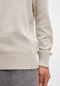 Pulover bej tricotat, cu un material texturat, manșete din tricot și un mic eticheta a brandului pe lateral. Purtați peste pantaloni gri.