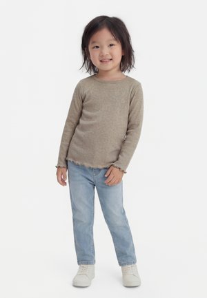 Jeune fille souriante debout, portant un haut beige à manches longues, un jean bleu clair et des baskets blanches sur un fond blanc uni.