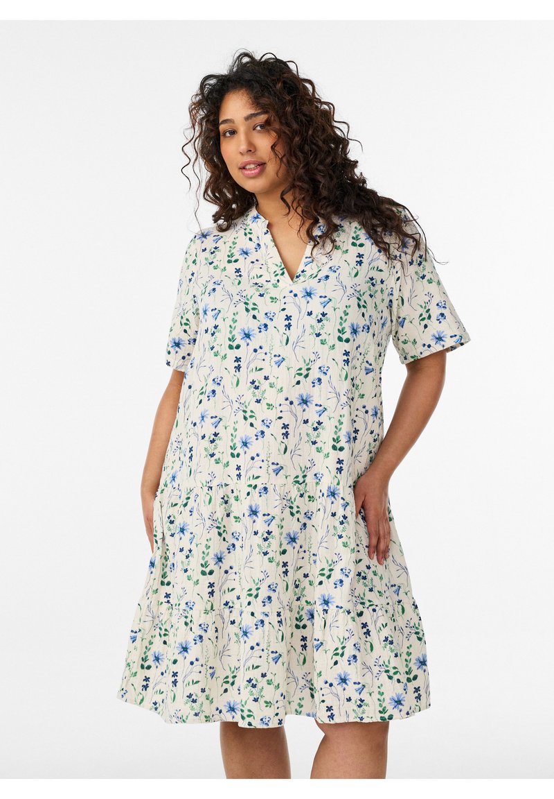 Moteris su ilgais garbanotais plaukais, dėvinti baltą, iki kelių siekiančią suknelę su mėlynais ir žaliais gėlių raštais, stovinti priešais paprastą foną.