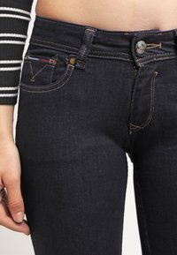 Jeans de mezclilla azul oscuro con un diseño ajustado, que cuentan con un pequeño bolsillo frontal, cierre de botón y costuras doradas en contraste.