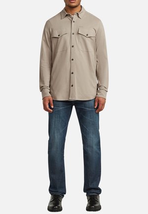 Homme portant une chemise beige à boutons avec poches poitrine, jean bleu et chaussures noires à lacets, debout devant un fond uni.