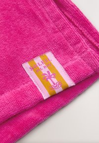 Asciugamano piegato di colore rosa brillante con un'etichetta a strisce che presenta un logo di una palma e il testo "Soleil All Day" nei colori rosa e giallo.