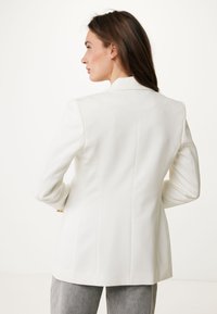 Bílý blazer s ostrými klopami, strukturovaným střihem a hladkým povrchem. Zadní pohled zobrazuje dobře padnoucí siluetu a dvě zadní švy.