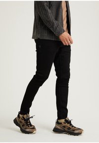 Jeans negros de corte ajustado, combinados con una camisa de cuadros gris y una camiseta beige, complementados con zapatillas marrones y negras con acentos texturizados.