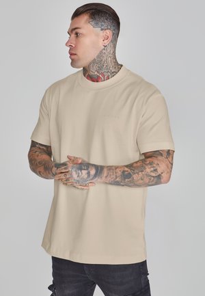 Beige, rövid ujjú póló, kerek nyakkivágással, puha anyagból, finom logó részlettel. A modell a ruházatot állva mutatja be.