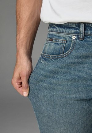 Blue denim jeans with a relaxed fit. Features include visible stitching, a front pocket, and a stainless steel button closure.