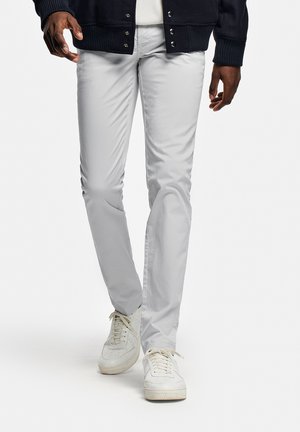 Personne portant un pantalon gris clair ajusté, des baskets blanches et un pull boutonné bleu marine foncé, marchant devant un fond blanc uni.