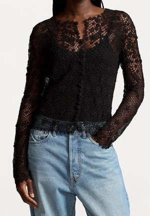 Blouse en dentelle noire à manches longues avec motifs floraux, boutonnée sur le devant, portée avec un jean denim bleu clair, une main dans la poche.