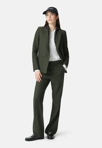 Donna in piedi che indossa un abito verde scuro con pantaloni a gamba larga, una maglietta bianca, un cappellino da baseball nero e mocassini neri, davanti a uno sfondo bianco uniforme.