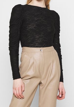 Kvinde iført en sort tekstureret bluse med lange ærmer og puffede skuldre samt beige, højtaljede plisserede bukser.