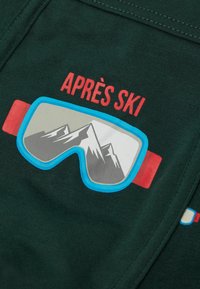 Grafika gogli narciarskich z odbiciem gór i czerwoną opaską na ciemnym tle, czerwony napis "APRES SKI" nad goglami.