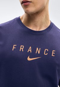 Κοντινό πλάνο ενός ατόμου που φοράει μπλε ναυτικό T-shirt με την επιγραφή «FRANCE» και το πορτοκαλί λογότυπο Nike στο στήθος.