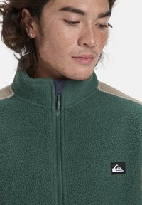 Grüne Fleece-Jacke mit hohem Kragen, Reißverschluss und tanfarbenen Schulterakzenten. Verfügt über ein kleines schwarzes Logo auf der Brust. Weiche Textur.