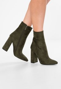 Bottines en daim vert olive, dotées d'un bout pointu, d'une fermeture éclair sur le côté et d'un talon épais. Texture lisse avec une silhouette élégante.