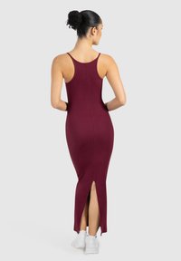 Kobieta z ciemnymi włosami upiętymi w kok, ubrana w bordową, bezrękawną sukienkę midi z tylnym rozcięciem i białe trampki, stojąca na jednolitym tle.