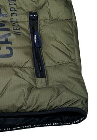 Wypchana oliwkowozielona kurtka z kieszenią na zamek i ciemnoniebieskimi akcentami. Posiada prążkowaną teksturę i branding wzdłuż dołu.