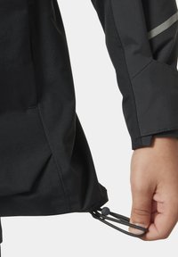 Czarna kurtka o gładkiej teksturze ma sznurki w dolnej części oraz szare odblaskowe akcenty na rękawie.