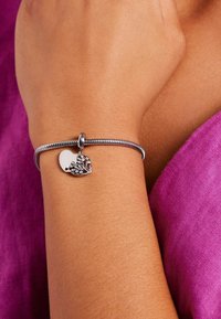 Zilveren armband met een hartvormige bedel met een bloemenpatroon. De armband heeft een gladde, flexibele textuur en wordt gedragen om een gebruinde pols.