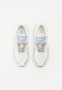 Chaussures de sport blanches et bleu clair en mesh et matériau synthétique, avec des lacets gris et une languette rembourrée. Logo "N" distinctif sur les côtés.