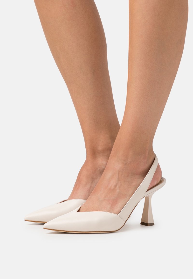 Jambes portant des escarpins blancs à bout pointu et bride arrière avec un talon incurvé et évasé sur un fond clair uni.