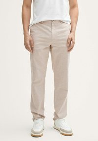 Chinos de color beige claro hechos de una tela texturizada y suave. Presentan una silueta clásica con cremallera y bolsillos delanteros, combinados con zapatillas blancas.