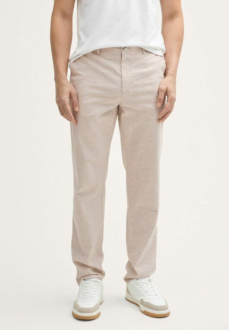 Chinos de color beige claro hechos de una tela texturizada y suave. Presentan una silueta clásica con cremallera y bolsillos delanteros, combinados con zapatillas blancas.