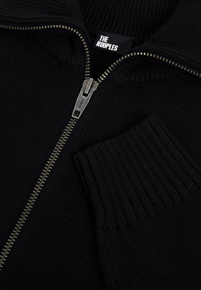 Pull en tricot noir côtelé avec une fermeture éclair argentée partiellement ouverte, montrant l'étiquette de la marque "The Kooples" et le tirette de fermeture éclair argentée assortie.