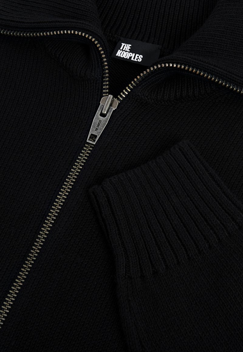 Schwarzer, gerippter Strickpullover mit silbernem Reißverschluss, der teilweise geöffnet ist und das Markenetikett "The Kooples" sowie den passenden silbernen Reißverschlussanhänger zeigt.
