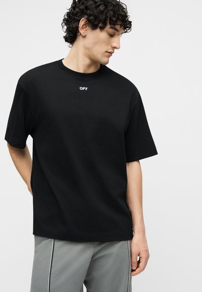 Νέος άνδρας με σγουρά μαλλιά, φορώντας μαύρο μπλουζάκι με την επιγραφή "OFF" και γκρι παντελόνι, κοιτάζοντας προς τα κάτω και πλάγια, μπροστά σε απλό φόντο.