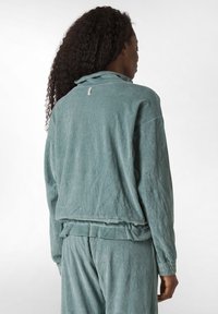 Jachetă din catifea de culoare teal, cu o croială lejeră, textură încrețită și tiv elastic. Prezintă un guler înalt și o etichetă la gâtul din spate.