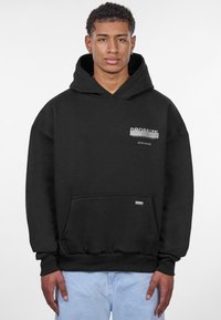 Sweatshirt à capuche noire avec une poche avant, arborant un logo et du texte imprimés en blanc sur la poitrine. Texture douce, coupe décontractée.