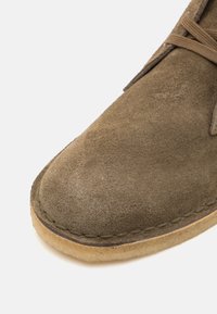 Ruskeaharmaat mokkanahkakengät, joissa on pyöreä kärki, beige kumi- ja näkyvällä ompeleella. Nauhat on sidottu yläosaan.