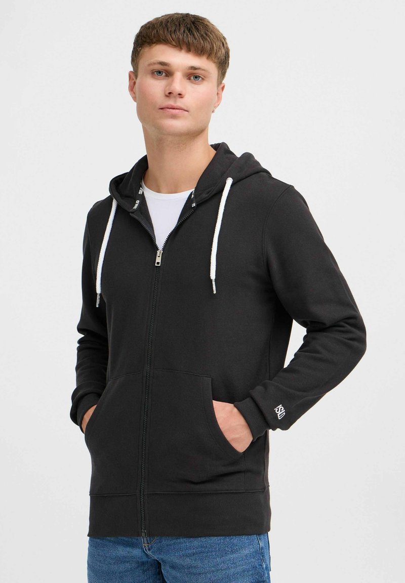 Sudadera negra con cremallera hecha de una tela suave, con cordones blancos, bolsillos tipo canguro y puños de canalé. Llevada sobre una camiseta blanca.