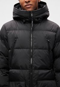 Casaco acolchoado preto com gola alta, fecho de correr frontal e dois bolsos no peito com zíper. Confeccionado em material liso e isolante com painéis costurados.