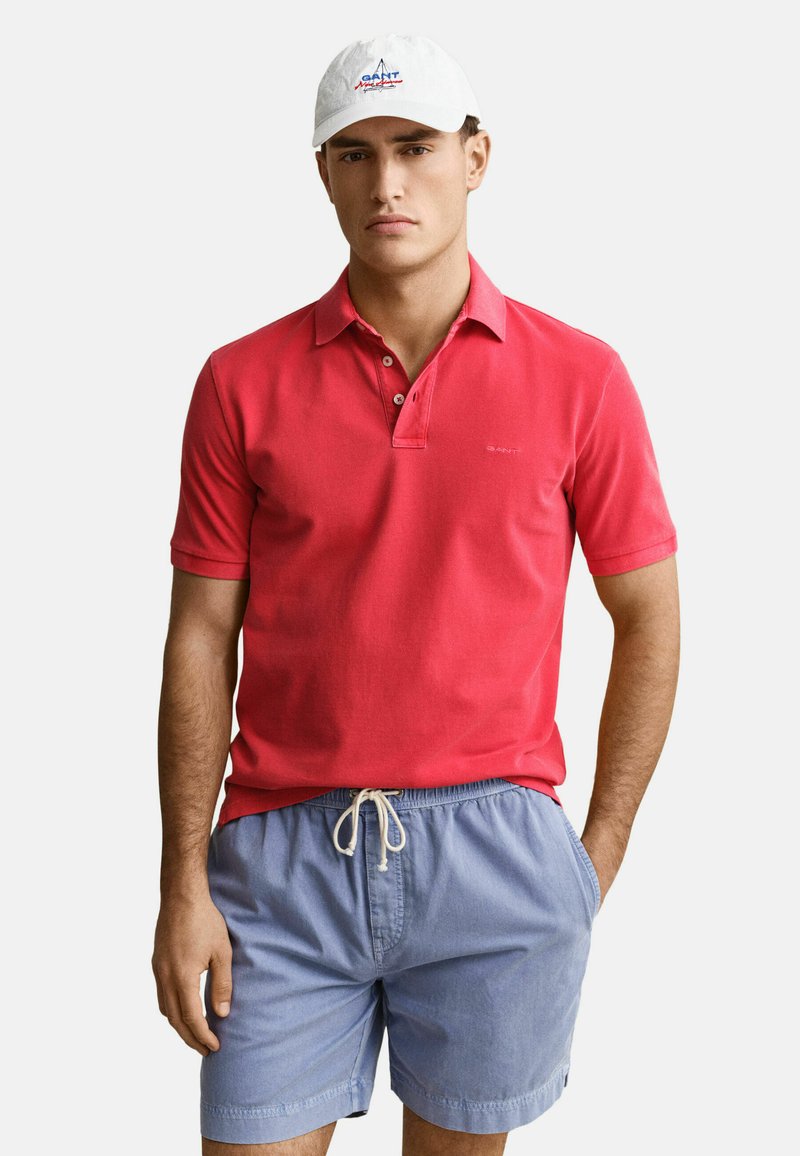 Mann mit weißer Kappe, leuchtend rotem Poloshirt und hellblauen Shorts mit Kordelzug, steht mit einer Hand in der Tasche vor weißem Hintergrund.