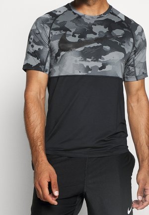 Mann trägt ein kurzärmeliges schwarzes und graues Tarnmuster-Nike-Shirt und schwarze Shorts mit Nike-Logo, steht mit entspannt an den Seiten hängenden Armen.