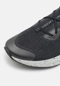 Pointe de chaussure de sport en mesh noir avec lacets noirs et texte "LACOSTE" gris sur la languette, avec une semelle intermédiaire blanche tachetée sur fond blanc.