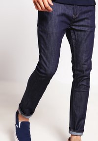 Mörkblå denimjeans med slim fit, som har en klassisk design, subtila sömmar och uppvikta mudd. Kombineras med marinblå slip-on skor.