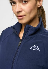 Marinblå fleecejacka med hög krage, dragkedja och Kappa-logotyp broderad i vitt på vänster sida. Mjuk textur och avslappnad design.