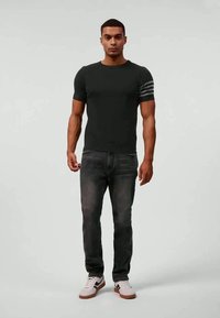Man wearing black short-sleeve shirt with three grey stripes on left arm, faded black jeans, and white sneakers with black and brown details.