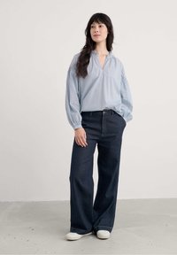 Frau trägt eine hellblau langärmlige Bluse, dunkle Weitbeinjeans und weiße Sneaker, steht mit einer Hand in der Tasche vor einem einfarbigen Hintergrund.