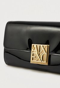 Μαύρο πατεντάριστο δερμάτινο τσαντάκι με ορθογώνια χρυσή κλειδαριά που διαθέτει κομμένες γραμματοσειρές, λεία υφή και προσεγμένα ραμμένα άκρα.