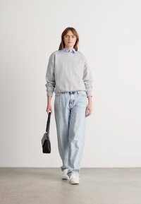Donna che cammina all'interno, indossando una felpa grigia, jeans azzurri chiari, sneakers bianche e portando una borsa nera nella mano destra.