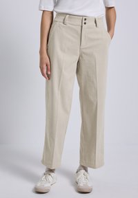 Vrouw in beige corduroy broek met dubbele knoopsluiting, witte sneakers, witte sokken en een wit shirt met korte mouwen, handen in de zakken.