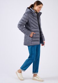 Giacca a vento grigia con cappuccio, design trapuntato, tasche laterali e zip frontale. Abbinata a jeans blu arrotolati e sneakers bianche.