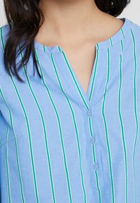 Chemise bleue à boutons avec des rayures verticales blanches et vertes, présentant un col en V et un tissu léger et texturé.