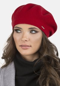 Red knit beret with a rounded shape, featuring subtle texture and a small metallic logo accent. Worn over a black turtleneck top.