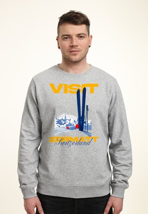 Man draagt een grijze sweater met een skisfeer van Zermatt, Zwitserland en het woord "VISIT" in grote gele letters.