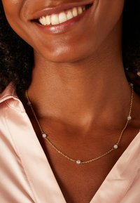 Gouden ketting met ronde lichtblauwe kralen, dicht bij het sleutelbeen gepositioneerd, gedragen met een satijnen roze shirt.